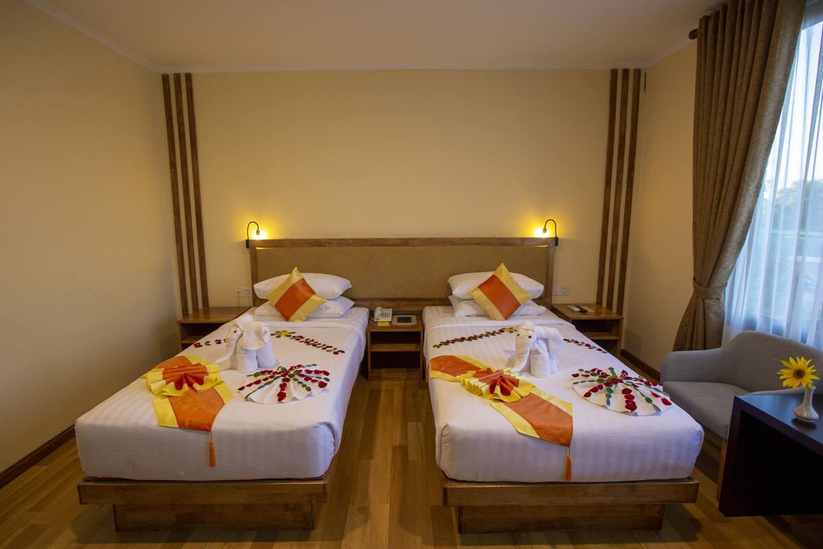 A hotel room with two single beds, wooden headboards, bedside tables with lamps, a small desk, and a chair.