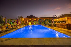 A large outdoor swimming pool with a thatched-roof structure on the right, surrounded by lounge chairs and umbrellas. - Acacia Hotel Bagan (Nyaung-U)