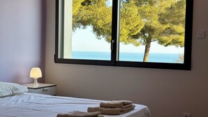 Un dormitorio con cama, mesita de noche, lámpara y ventana con vistas al mar y árboles.