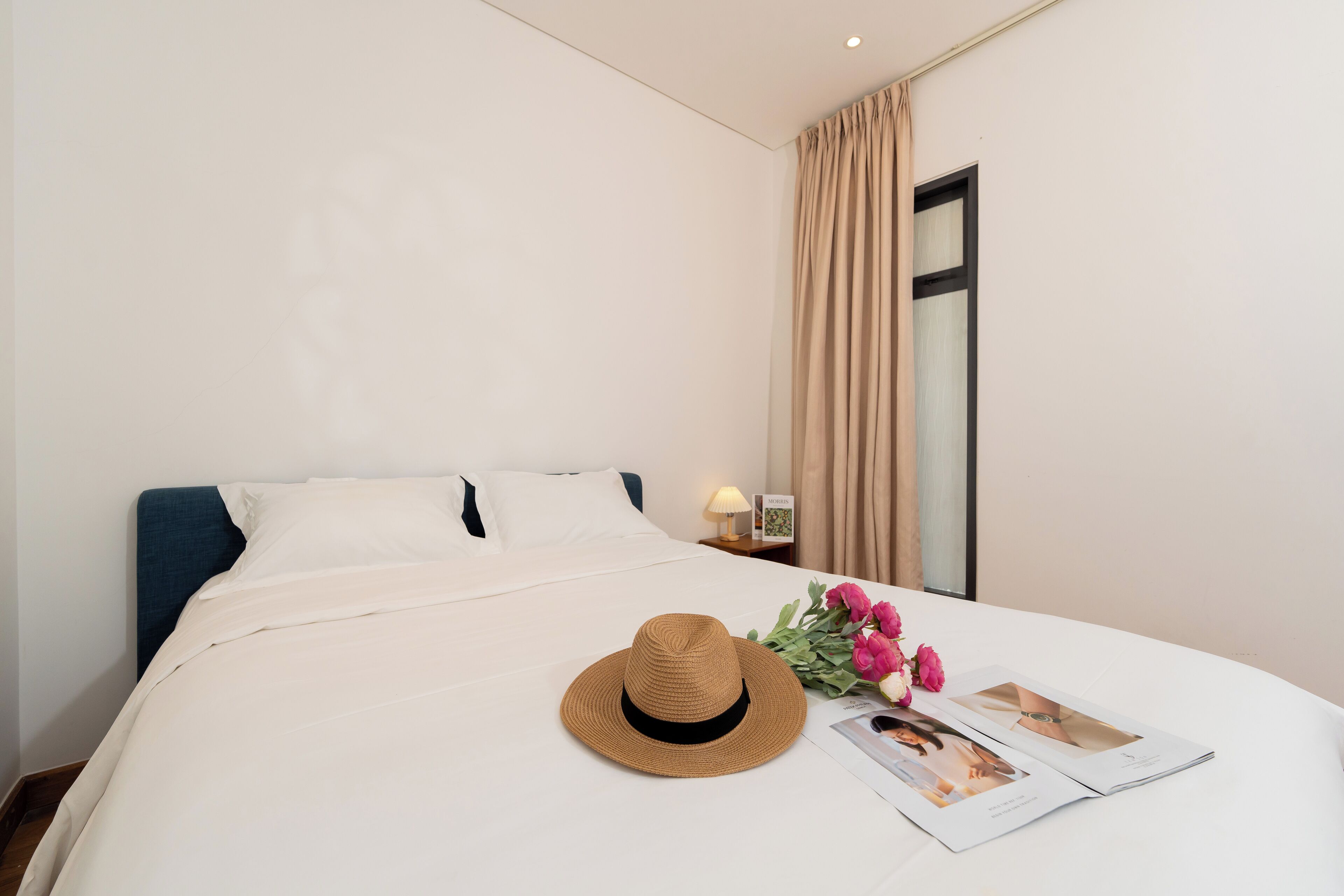 A neatly made bed with a white comforter, a brown hat, and a bouquet of pink flowers on the bed.