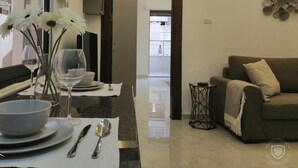A modern living room with a sofa, a dining table set with plates, glasses, and cutlery. There is a vase with flowers on the table, and a decorative wall piece above the sofa. - Amazing one Bedroom Apartment in Amman,elwebdah 1 (Amman)