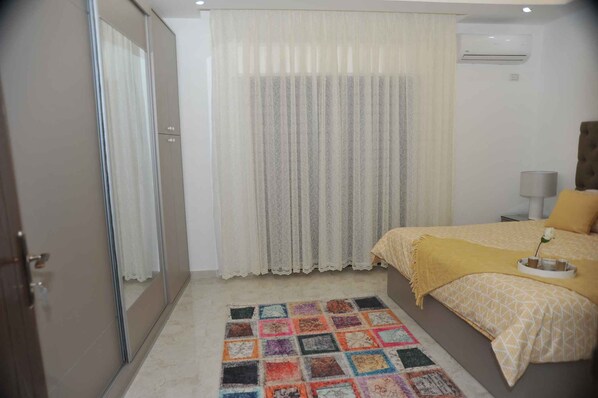 A bedroom with a bed, a bedside table, a lamp, a window with curtains, and a wall-mounted air conditioner. - Amazing one Bedroom Apartment in Amman,elwebdah 6 (Amman)