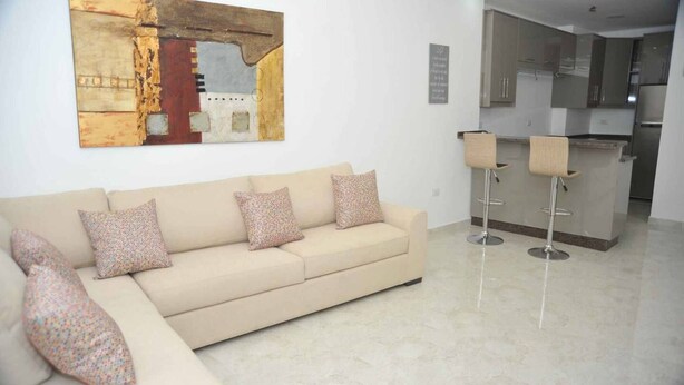 Un salon moderne avec un canapé beige, des coussins à motifs, une grande peinture abstraite et une cuisine en arrière-plan.