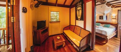 A cozy room with a wooden floor, a sofa, a TV, a small table, and a window with blinds.