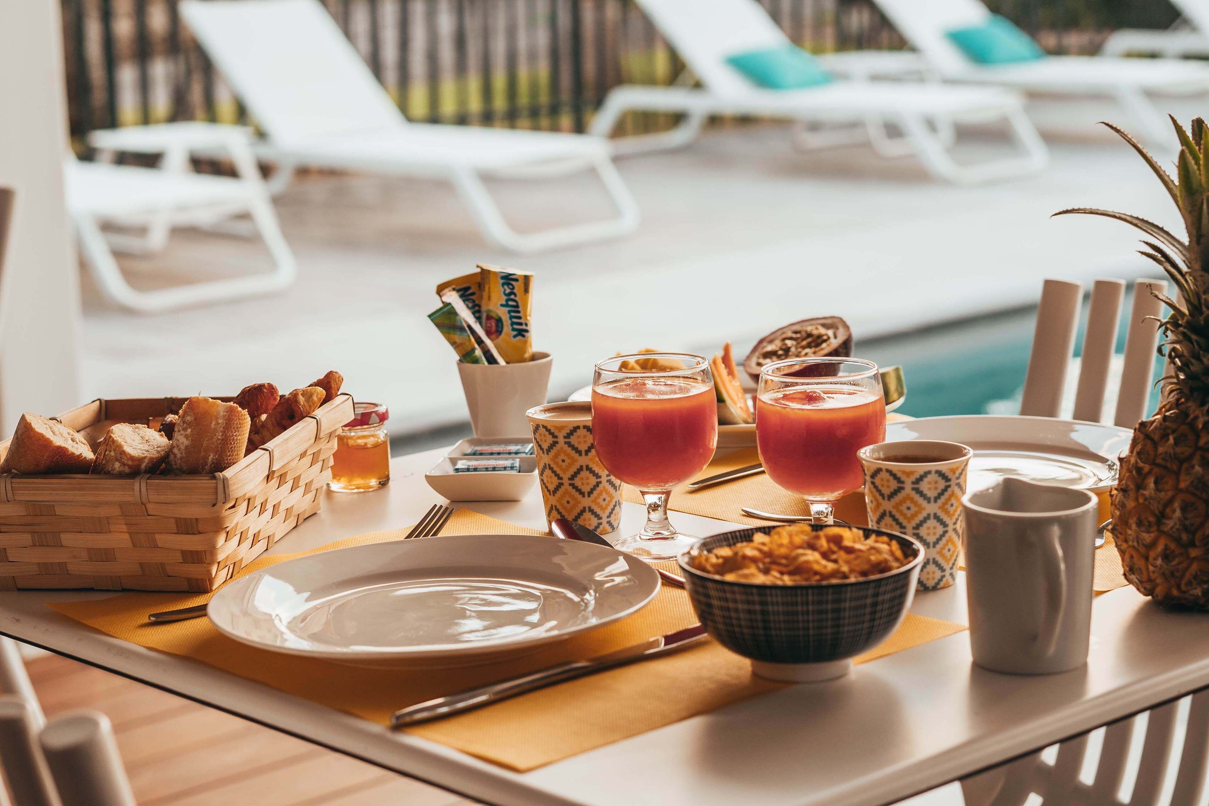 A breakfast setup by a poolside with a wicker basket of bread, a bowl of cereal, two glasses of orange juice, and a pineapple.