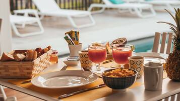 Disposição para o café da manhã junto a uma piscina, com uma cesta de vime de pão, uma tigela de cereais, dois copos de suco de laranja e uma abacaxi.