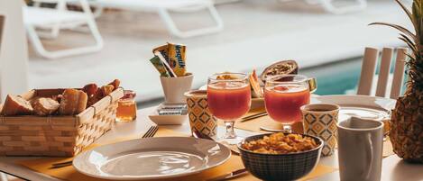 A breakfast setup by a poolside with a wicker basket of bread, a bowl of cereal, two glasses of orange juice, and a pineapple.