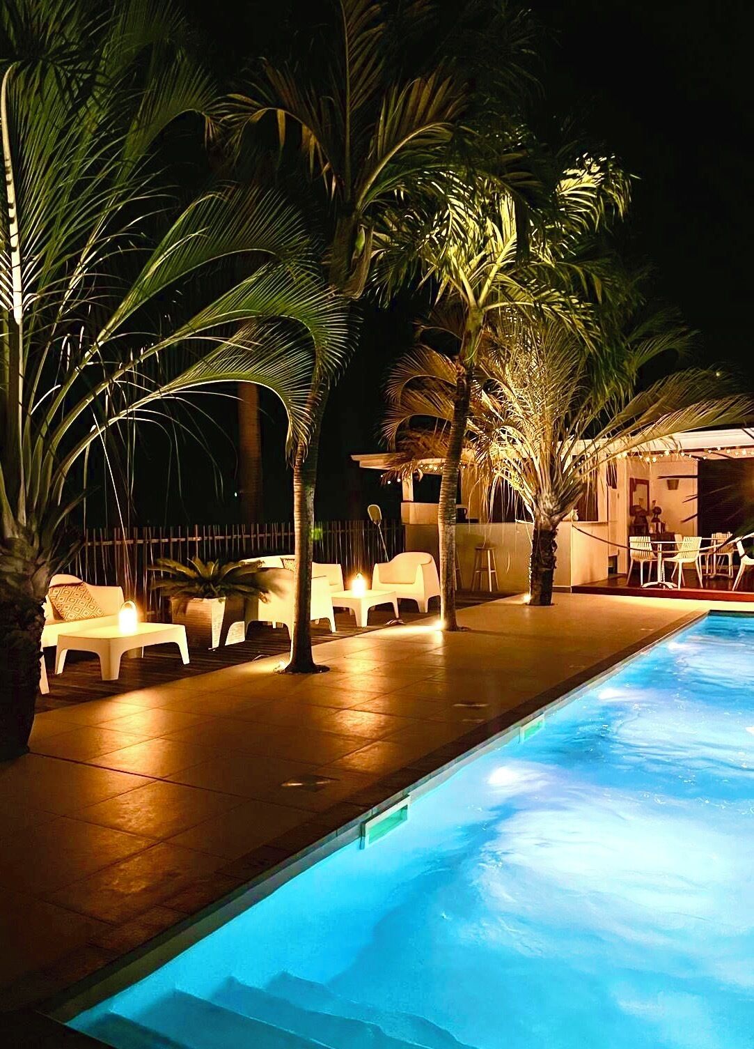 Zona de piscina nocturna con palmeras, sillas de descanso y un edificio al fondo.