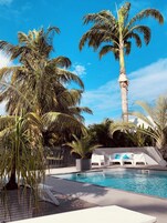 Área de piscina com palmeiras, cadeiras de lounge e uma parede branca com almofadas azuis.