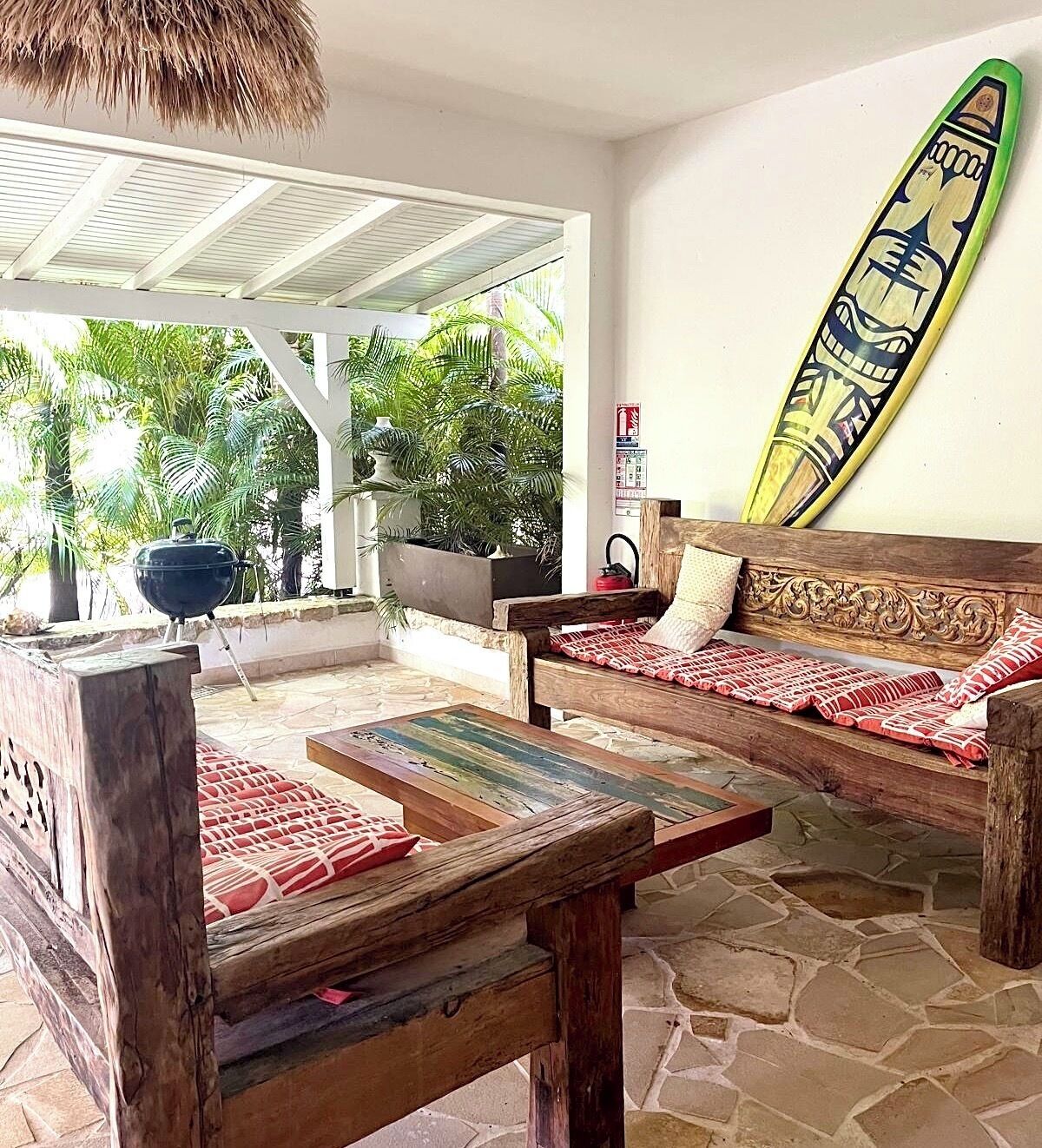 A wooden bench with a patterned cushion, a surfboard leaning against a wall, and a grill outside.
