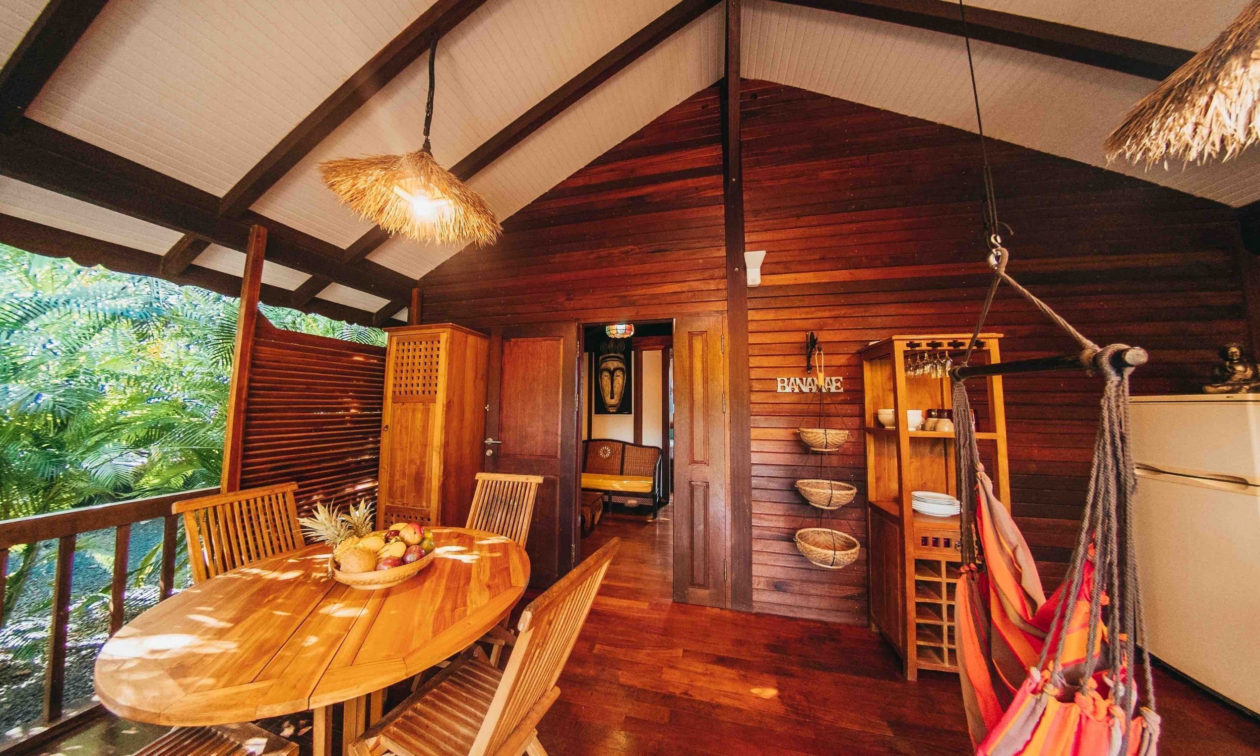 A wooden interior with a round wooden table, chairs, a hammock, and a wooden cabinet with a woven basket.