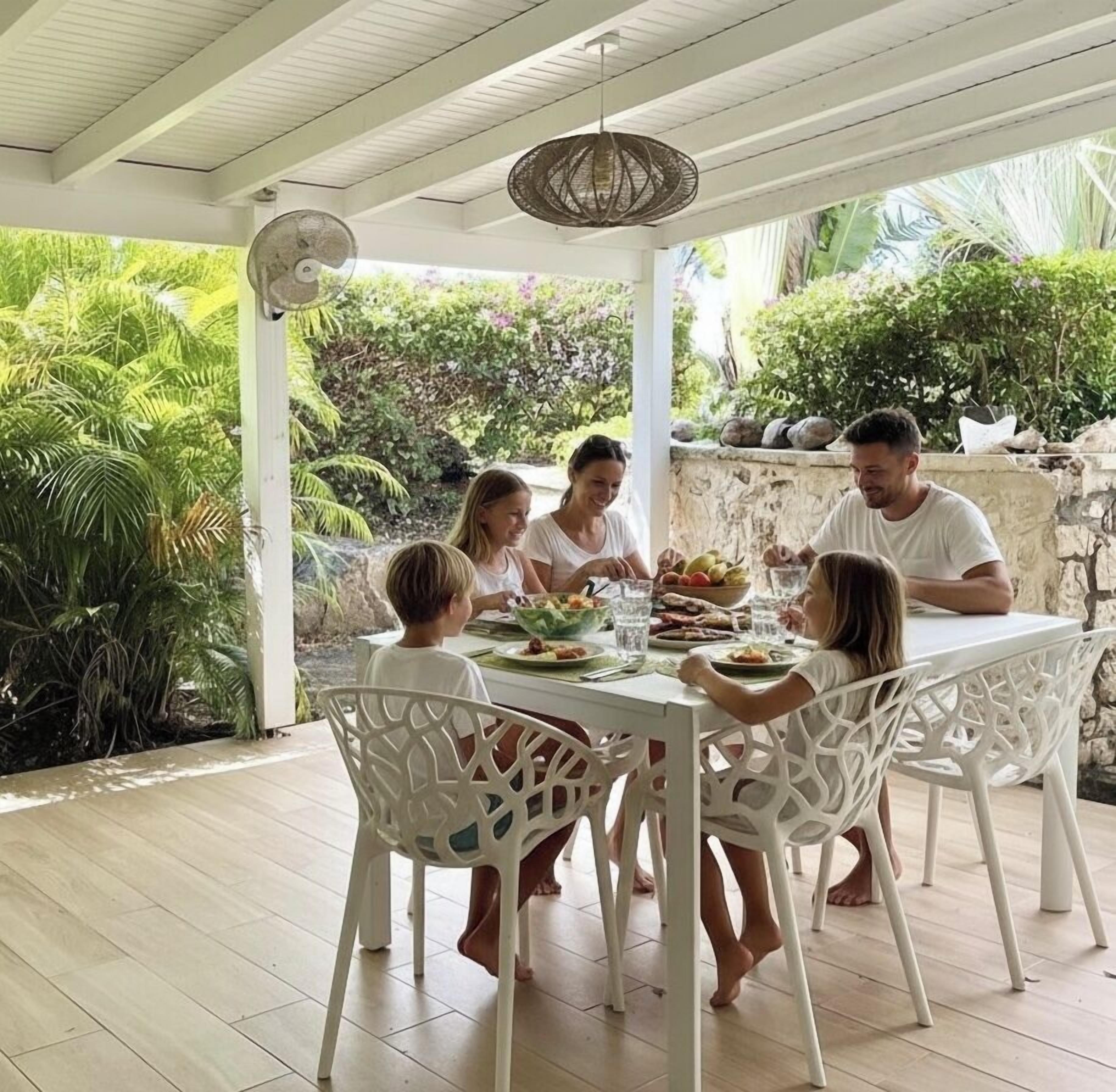 A family dining at a white table with chairs, surrounded by lush greenery and a stone wall.
