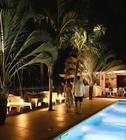 Área de piscina à noite, com palmeiras, espreguiçadeiras e piscina.