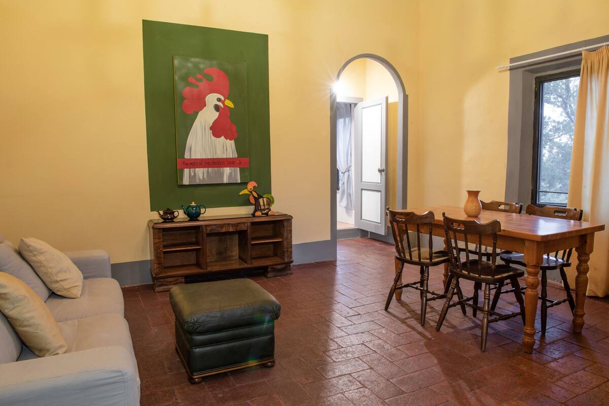 A dining area with a wooden table and chairs, a green wall with a rooster artwork, a sofa, and an ottoman.