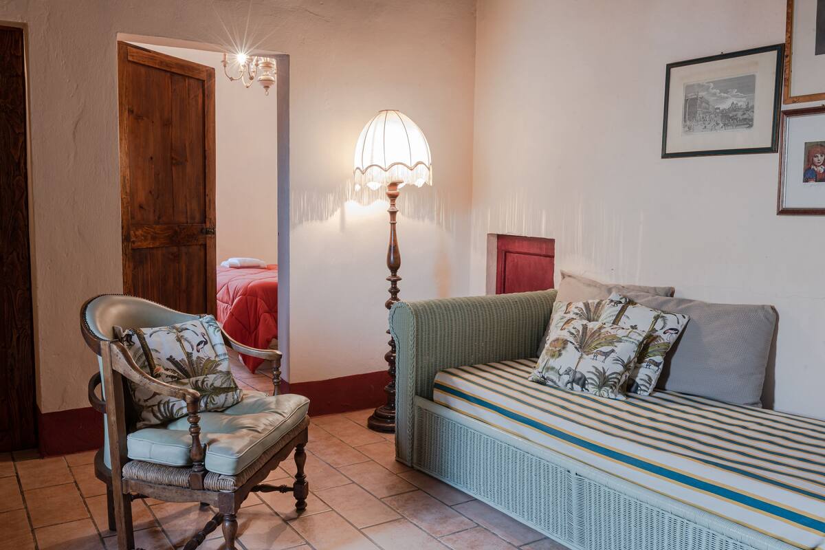 A room with a wooden door, a chair with a floral cushion, a sofa with striped cushions, a floor lamp, and a framed picture on the wall.