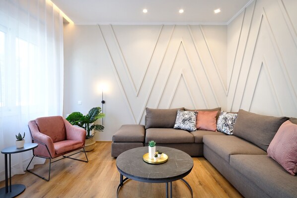 A modern living room with a sectional sofa, a pink armchair, a round coffee table, and a potted plant. - The Flirty Athens (Kallithea)