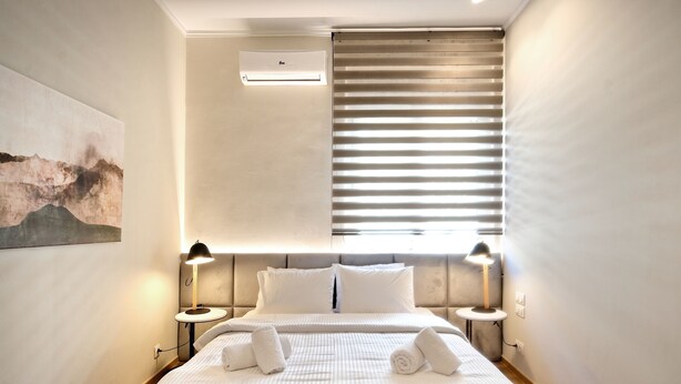 Una habitación de hotel moderna con una cama grande, dos mesitas de noche con lámparas, aire acondicionado en el techo y un ventanal con persianas.