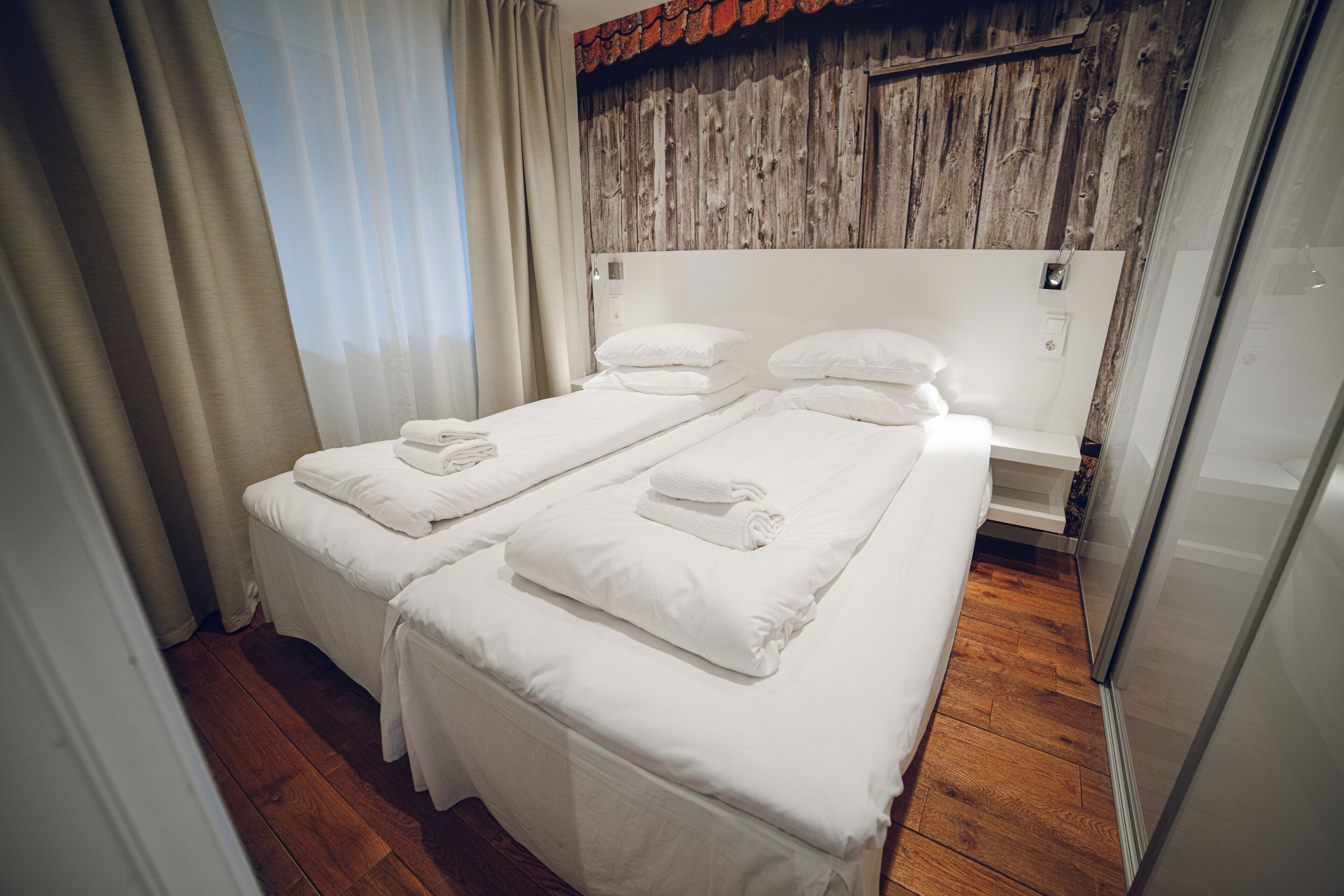 A hotel room with two single beds, wooden flooring, a wooden headboard, and a window with curtains.