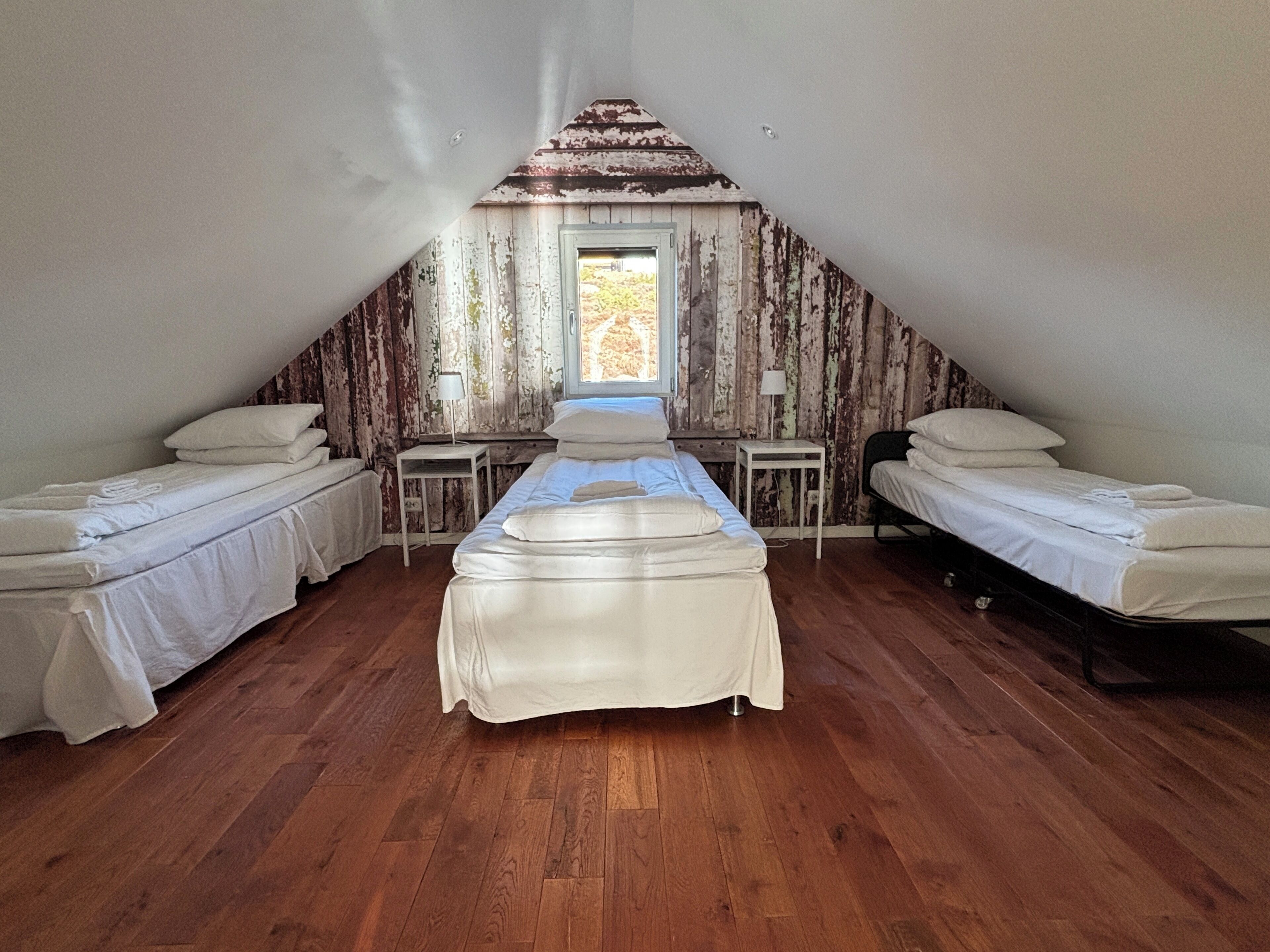 Quarto com duas camas de solteiro, uma cama maior no centro, pisos de madeira e teto inclinado com painéis de madeira.