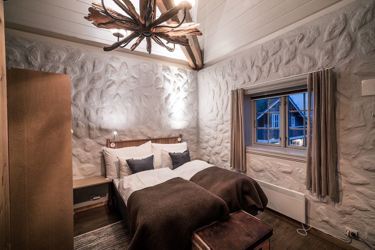 A bedroom with a rustic stone wall, a wooden ceiling, a large window with curtains, a bed with two pillows, and a nightstand.