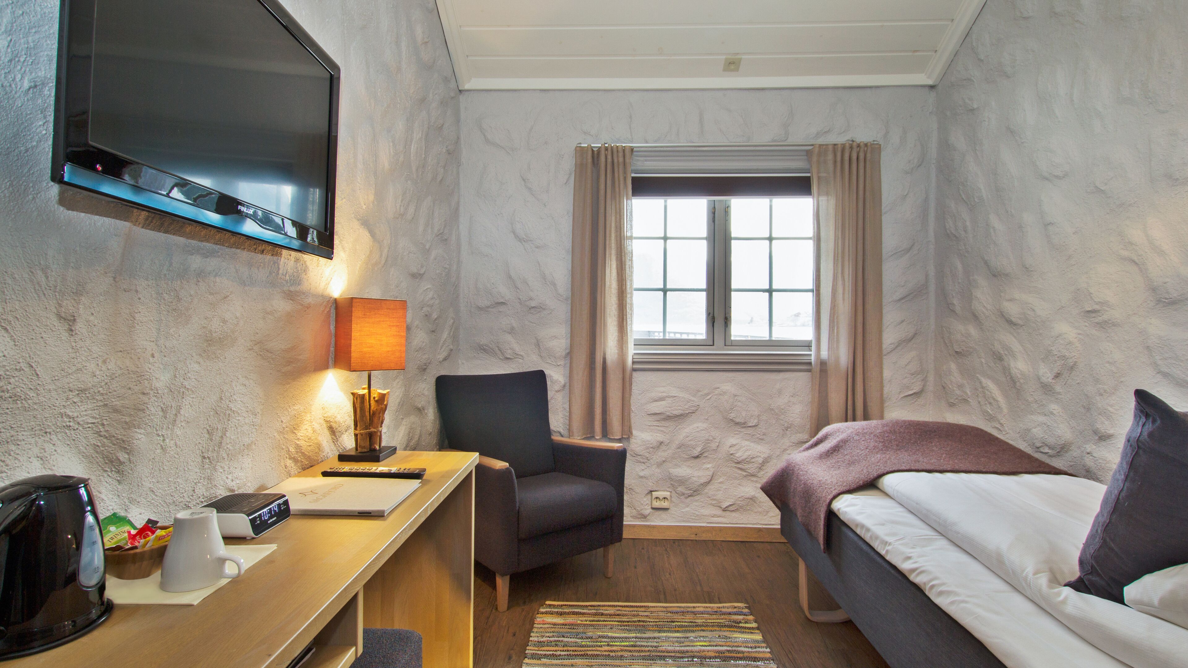 Ein Hotelzimmer mit Bett, Schreibtisch mit Lampe, Stuhl, Fernseher und einem Fenster mit Vorhängen.