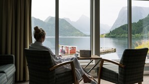 Quarto com uma grande janela com vista para um lago e montanhas, um laptop sobre uma mesinha e duas cadeiras.
