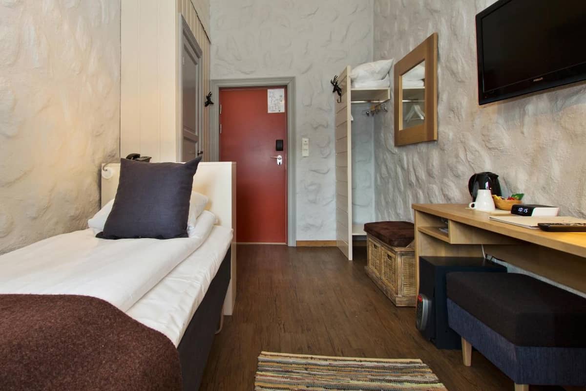 A compact hotel room with a single bed, a desk with a chair, a television mounted on the wall, and a small seating area with a bench.