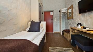 Habitación de hotel compacta con una cama individual, un escritorio con silla, un televisor montado en la pared y una pequeña zona de estar con un banco.