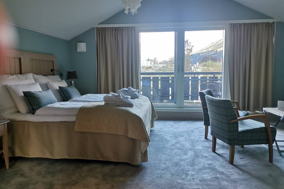 A bedroom with a large bed, a desk, and a chair. There is a window with curtains and a view of the outdoors.
