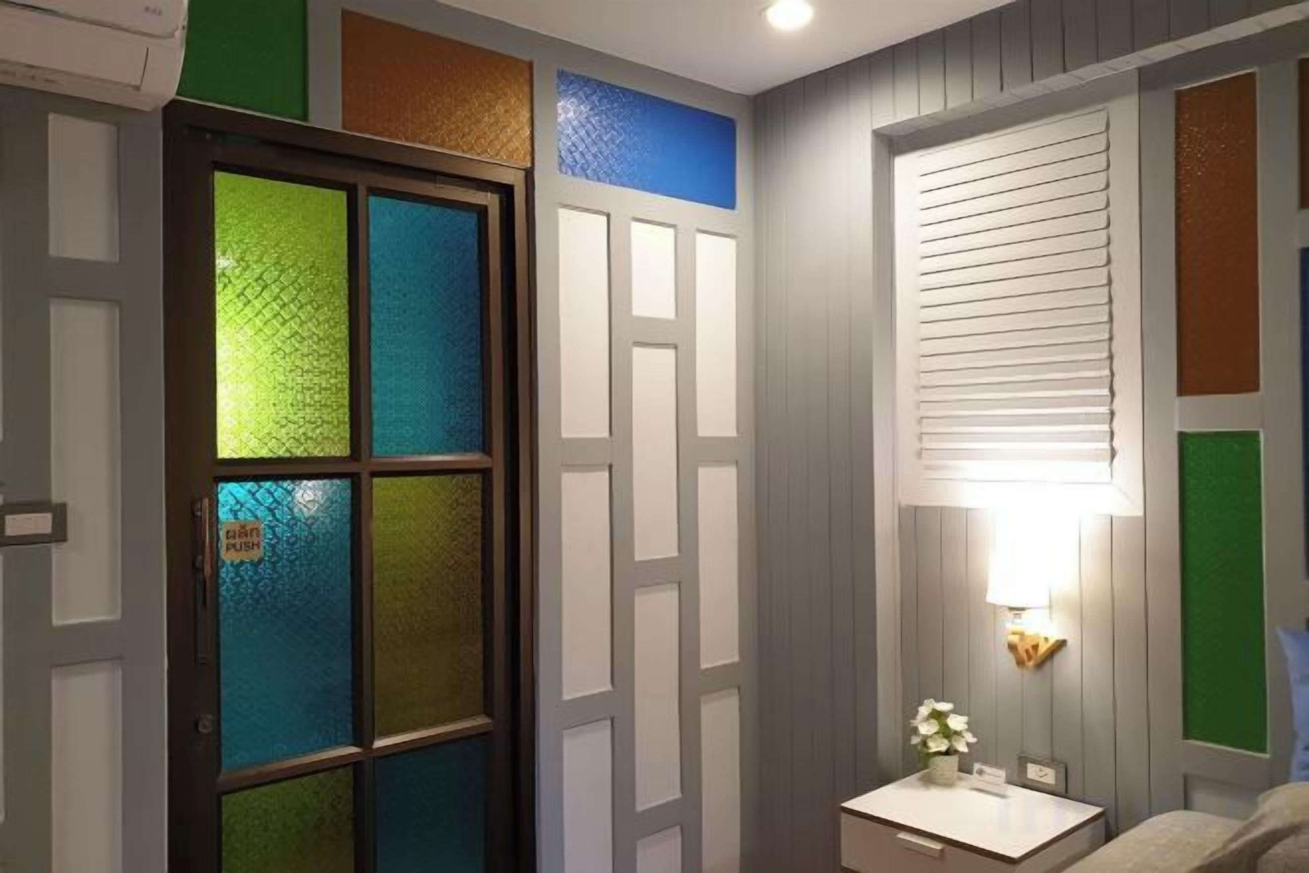 A room with a door featuring colored glass panels, a wall-mounted light fixture, and a small bedside table with a flower.