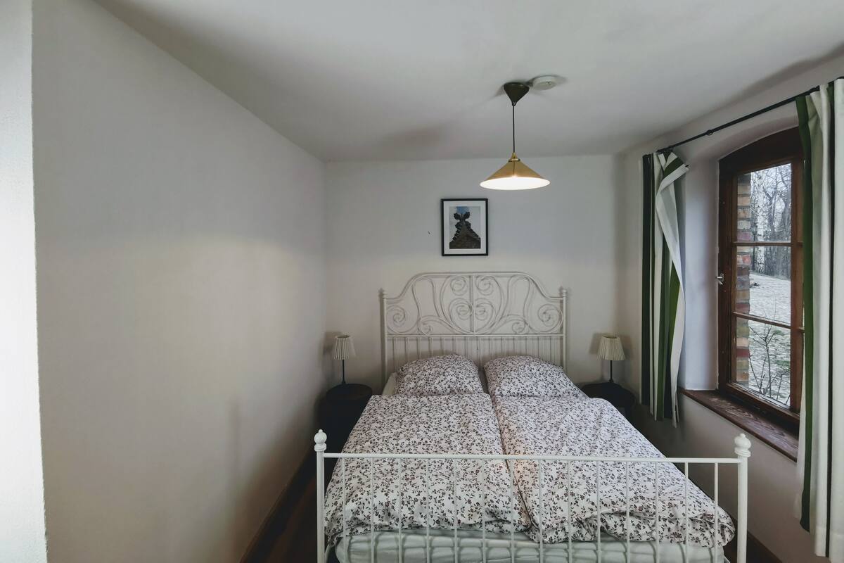 Quarto com cama de metal branca, uma mesa de cabeceira individual com abajur, luminária no teto, quadro na parede e janela com cortinas.