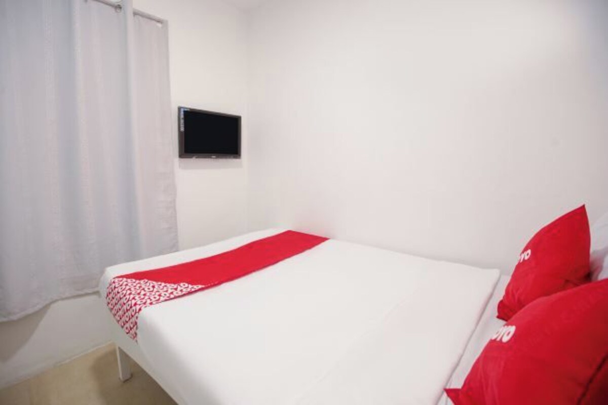 A single bed with a red and white bedspread, a black television mounted on the wall, and a white curtain.