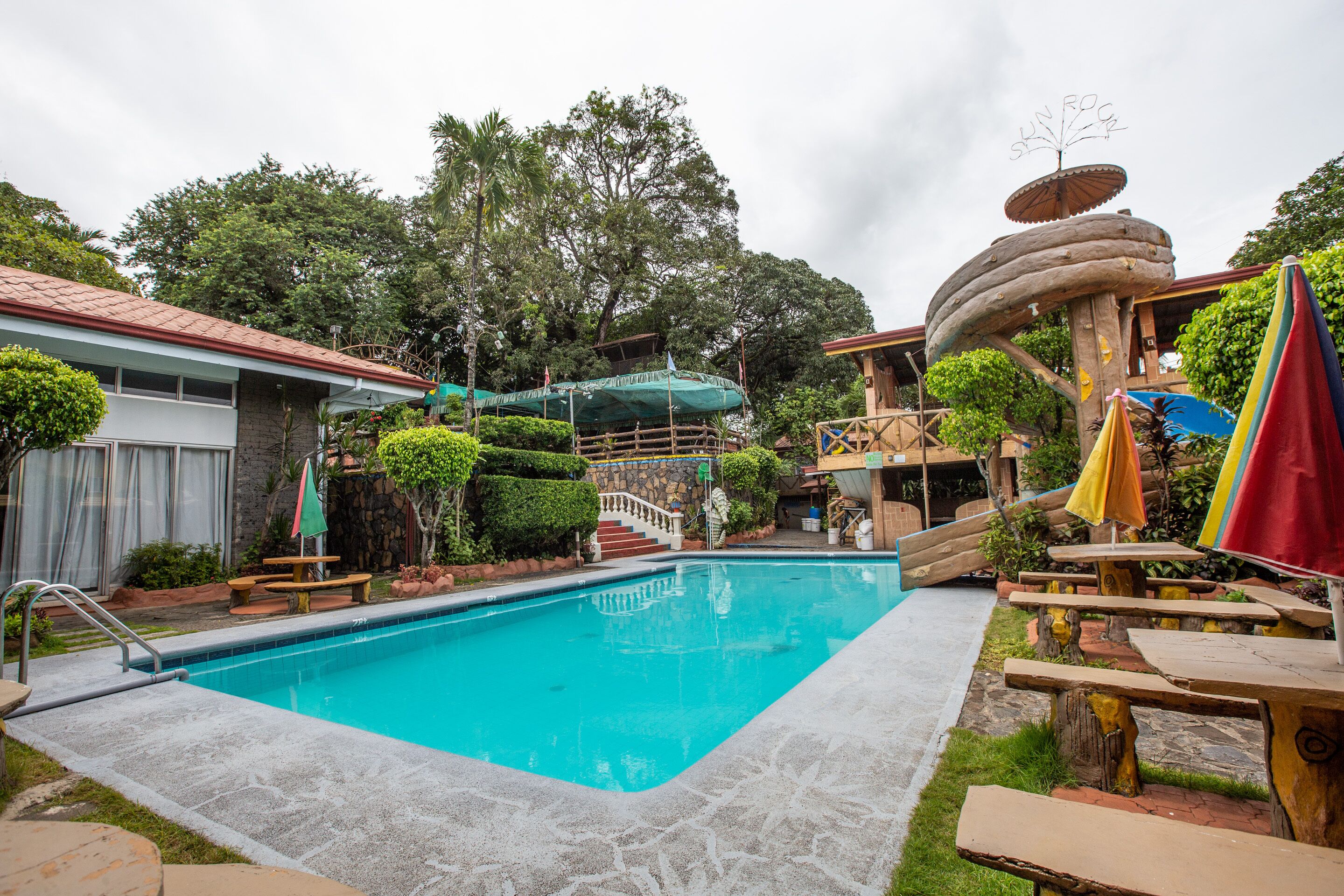 Una piscina con mesas de picnic y sombrillas, rodeada de follaje y un edificio de piedra.