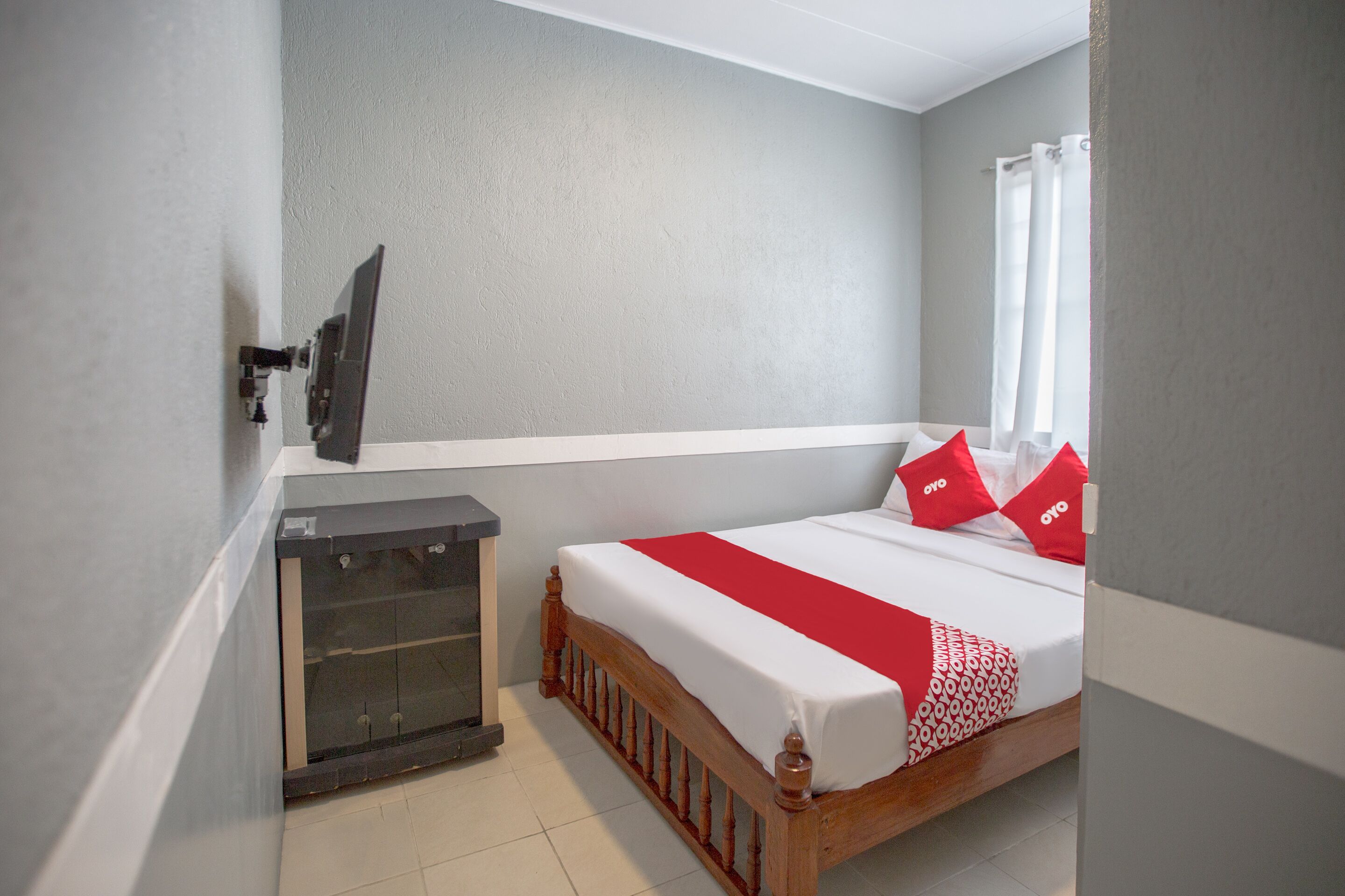 A bedroom with a wooden bed, white and red bedding, a TV mounted on the wall, and a small cabinet.