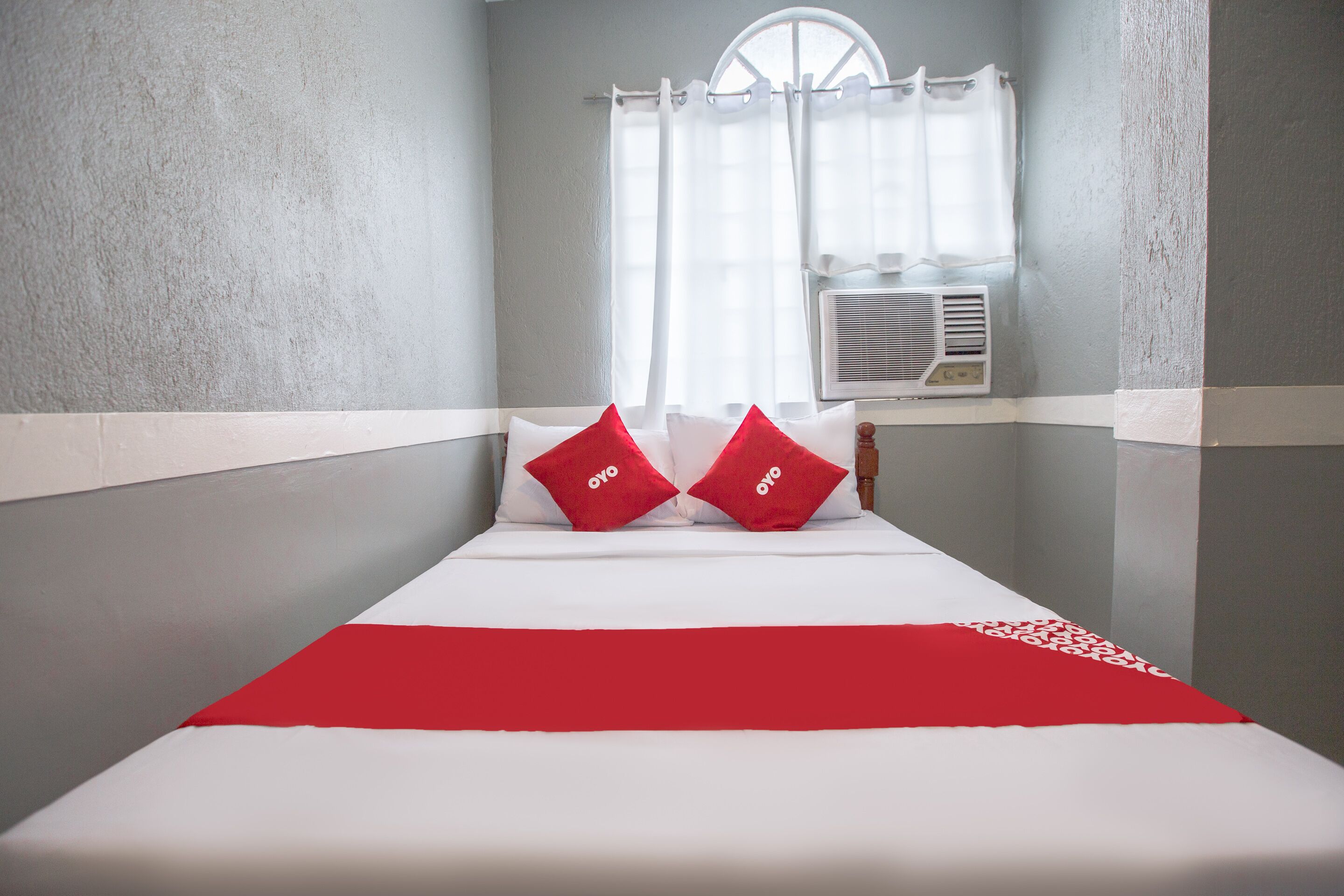 Habitación de hotel con una cama, dos almohadas rojas, una colcha blanca y una ventana con cortina blanca.