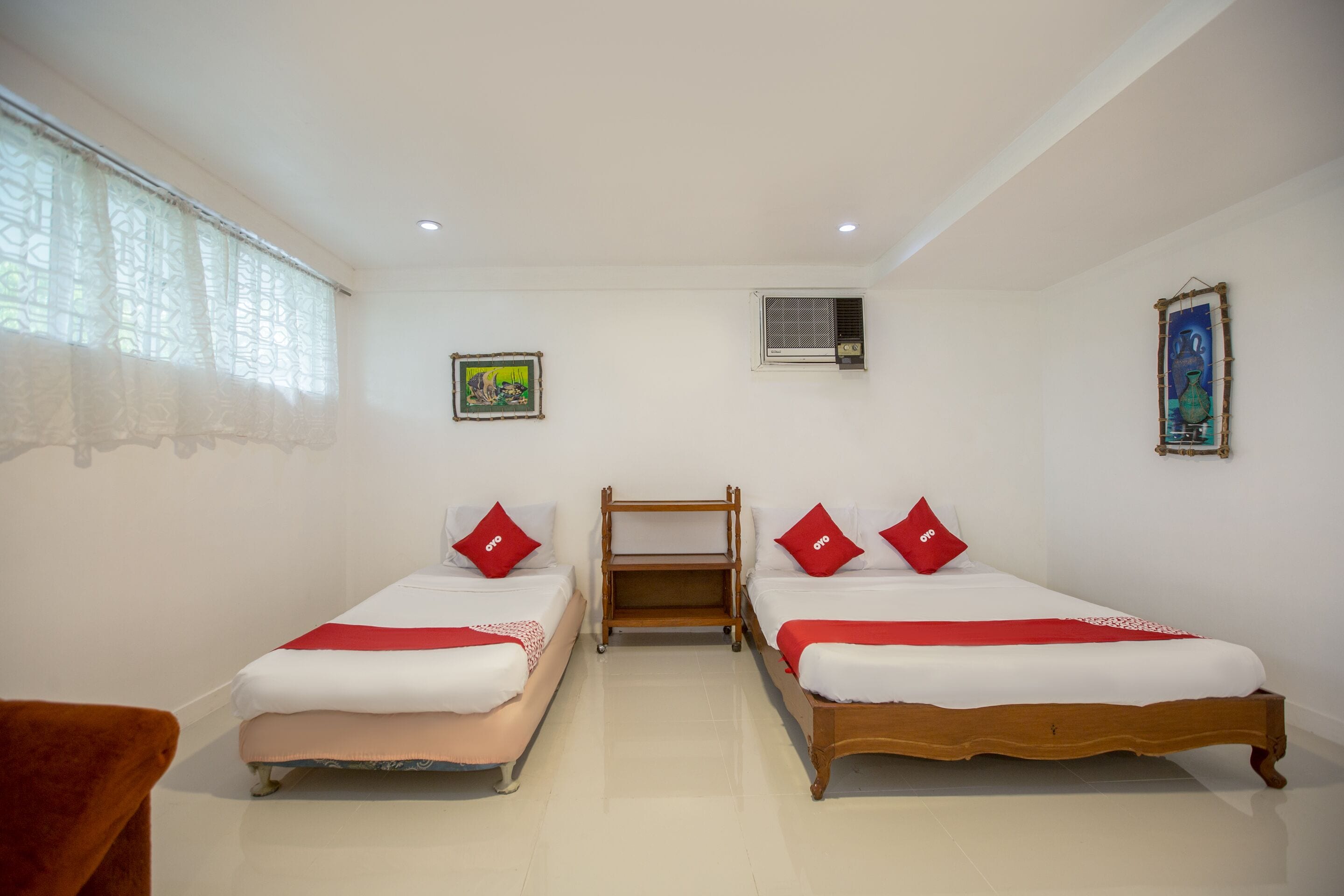 Habitación de hotel con dos camas individuales, una mesita de noche de madera, una pequeña mesa y una ventana con cortinas de gasa.