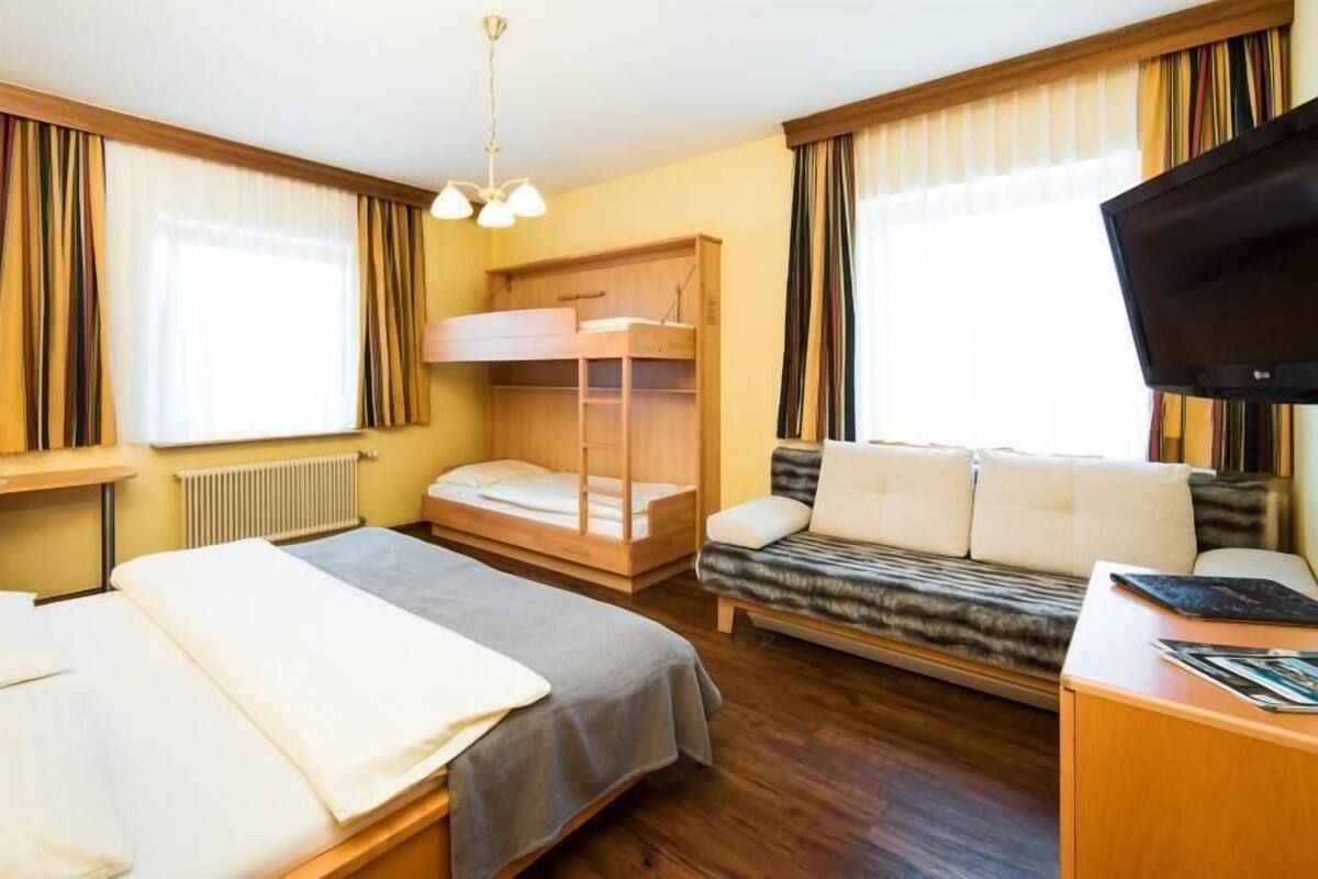A hotel room with a bunk bed, a sofa, a desk with a flat-screen TV, and a window with curtains.