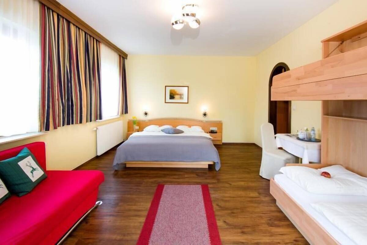 A room with a bed, a red sofa, a wooden bunk bed, a small table, and a window with curtains.