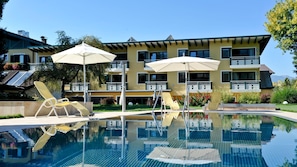 A swimming pool with lounge chairs and umbrellas, a multi-story building with balconies, and a clear sky. - Familiengut Ertlhof (Seeboden)