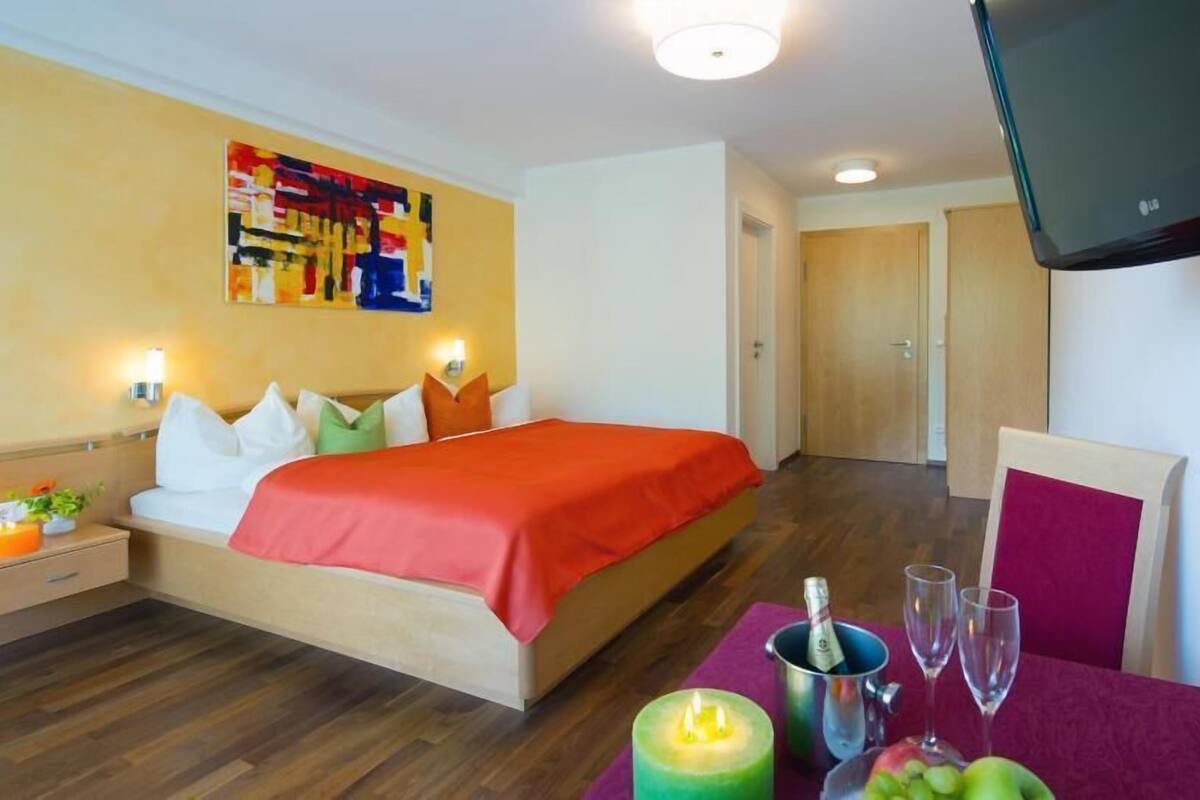 A hotel room with a bed, bedside table, wall art, TV, and a table with a bottle, glasses, and fruit.