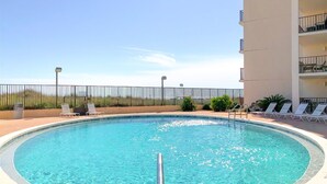 A clear blue pool with a metal ladder, surrounded by a low brick wall and a metal fence. There are lounge chairs and a building with balconies in the background.