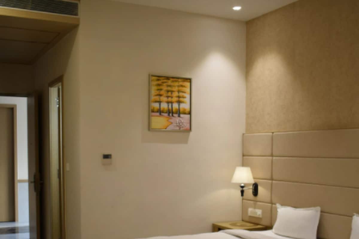 A hotel room with a large bed, a nightstand with a lamp, a painting on the wall, and a door leading to another room.