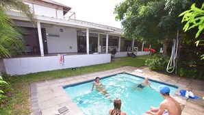 A swimming pool with a covered patio area, a grassy yard, and a house in the background. - Baleia Azul 26 (Zitundo)