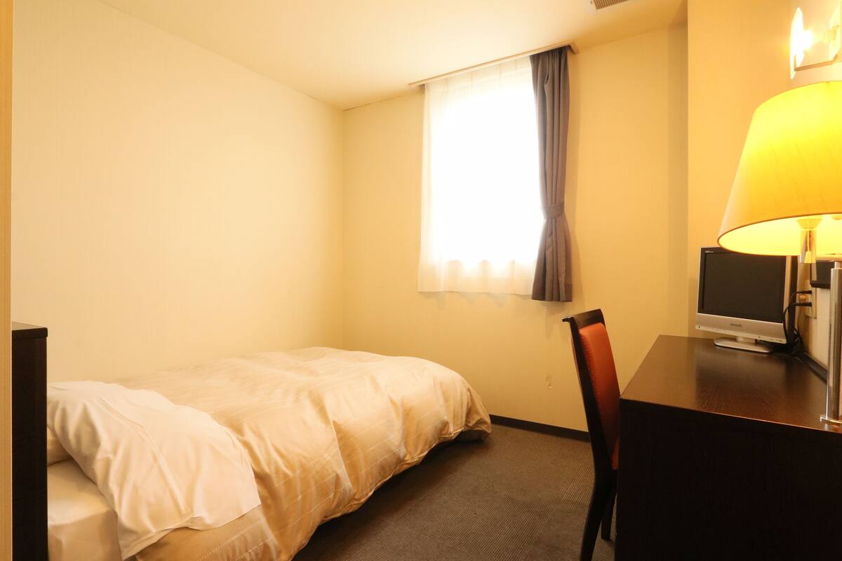 A hotel room with a bed, a desk with a lamp, a television, and a window with curtains.