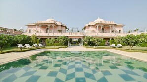 Un edificio grande con dos cúpulas, un patio central con piscina a cuadros y varias sillas de descanso.