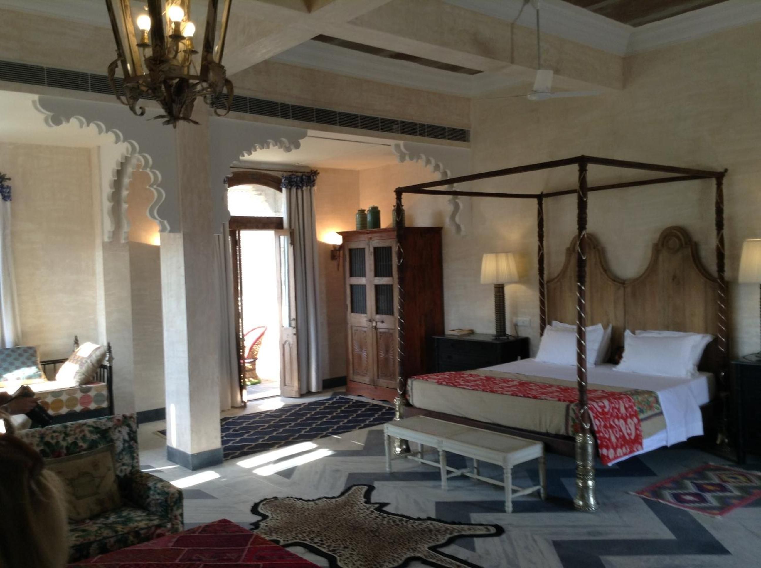 A four-poster bed with a red and white bedspread, a wooden nightstand, a lamp, and a chair with a floral pattern.