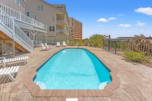 A rectangular swimming pool with a wooden deck surrounding it, a covered seating area with chairs, and a building with multiple balconies in the background.