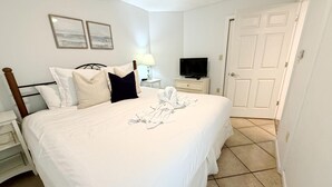A neatly made bed with white bedding and a few pillows, a bedside table with a lamp and a television, and a door in the background. - Moondrifter 105 (Panama City Beach)