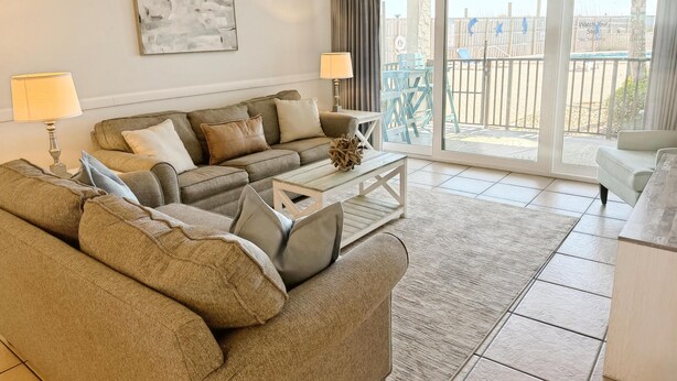 A living room with a sofa, a coffee table, and a view of a balcony with outdoor furniture. - Moondrifter 105 (Panama City Beach)