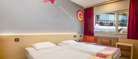 Habitación de hotel con dos camas, un escritorio, un ventanal con cortinas rojas y una pared con un diseño circular y texto.